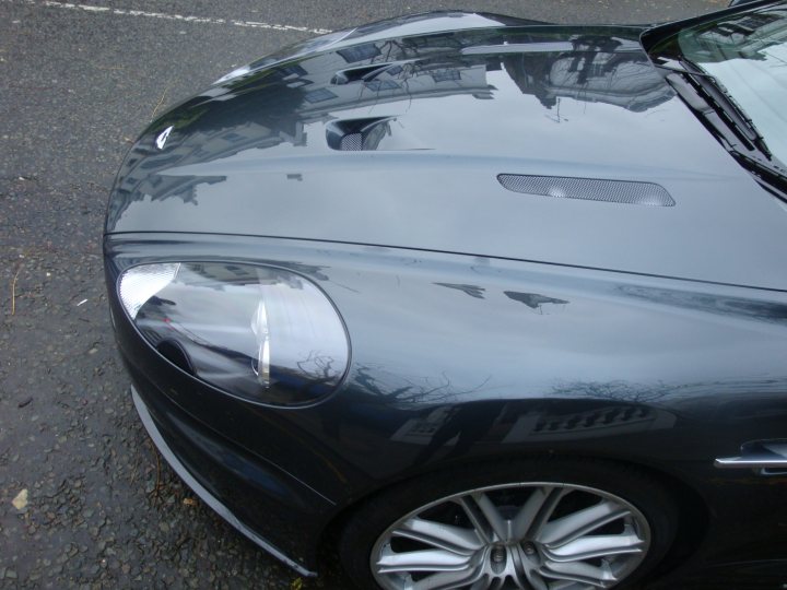 Astons Tempest Blue Pistonheads - The image showcases a black car parked on a wet street, as revealed by the sheen on the pavement. The car has sleek curves and a shiny paint job that catches the light. On the right side of the image, the white, textured fabric of the car's hood stretches out into the expanse of the dark gray sky, dotted with scattered clouds. On the left, the silver-colored BMW logo proudly displays its presence on the hood. The contrast between the dark car and the white textured fabric is striking.