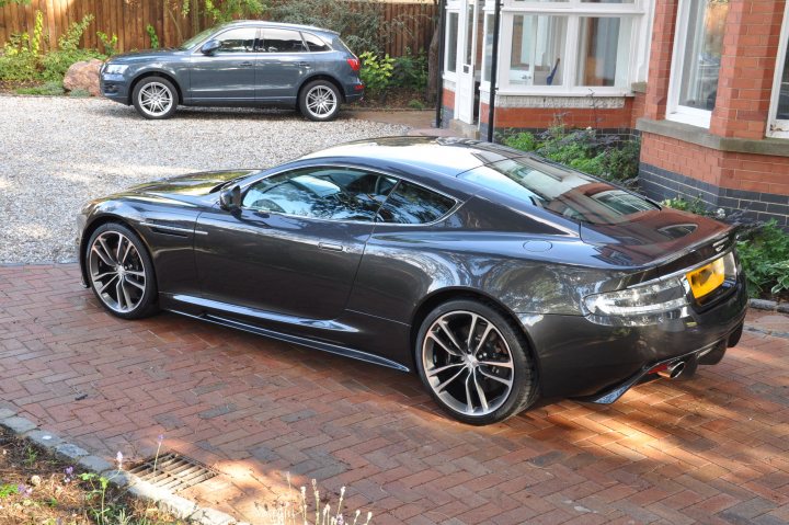 Finally Pistonheads Dbs Vantage - The image features a sleek black sports car parked on a driveway. The car's glossy finish reflects the surrounding environment, indicating it's a bright day. The car is positioned on the right side of the image, with its front facing the viewer. In the background, there's another car parked on the left, and a house is visible in the top right corner. The driveway appears to be made of brick pavers, adding a rustic charm to the scene. The clear sky in the background suggests good weather.