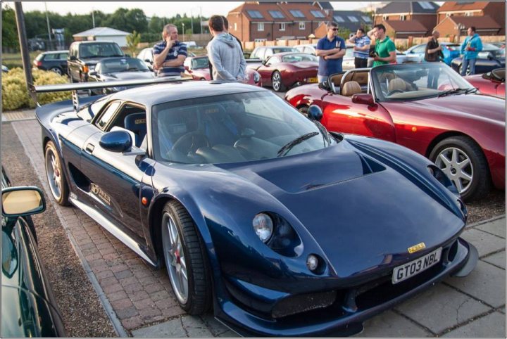 TVRCC + Guests Berkshire June Meet - Page 1 - Thames Valley & Surrey - PistonHeads - The image depicts a gathering of people and cars on a paved surface. The primary focus is a blue sports car, parked within the frame of the image. It's a sleek vehicle with aerodynamic features, and it occupies a significant portion of the scene. Surrounding this central car is a group of individuals standing close, seemingly engaged in conversation or admiring the vehicle. The background is populated with several other cars, suggesting the setting could be a car show or a similar event. The time of day appears to be either dusk or dawn, given the soft lighting that casts gentle shadows. The overall atmosphere of the image suggests a casual yet enthusiastic gathering, likely centered around the appreciation of automobiles.