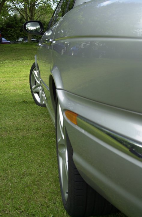 Pistonheads Super Sunday Dyvels Corbridge - The image shows the side profile of a silver car, which appears to be parked on the grass. The car has a shiny finish and features a large alloy wheel. The surrounding area seems to be a well-kept lawn or a garden, with lush green grass and a hint of trees in the background. The sky is partially visible, suggesting a bright day, and there are other vehicles and people in the background, indicating that this might be a public area. The perspective of the photo is from an angle that captures the car's wheel and part of its body, emphasizing its design and shine.