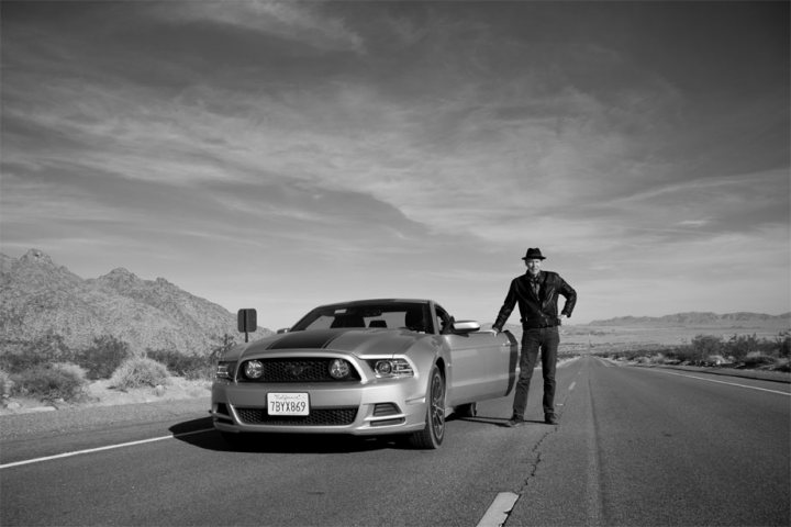 Advice please - Bullitt Mustang - Page 1 - Mustangs - PistonHeads - In the black and white image, a coasting, sleek muscle car is the center of attention. A man in a black jacket and hat stands confidently on the side of the road, leaning against the car with an air of nonchalance. The highway stretches out ahead of them, curving gradually in the open range landscape. Nearby, a fenced-off area suggests a boundary or a public roadside park, adding to the sense of adventure and freedom.