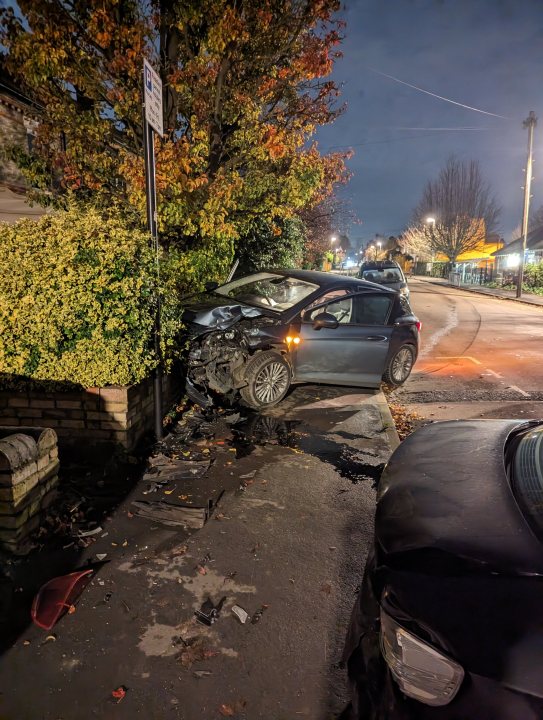 Someone crashed into my house and my car!  - Page 1 - General Gassing - PistonHeads UK - The image shows a nighttime scene of a car accident. A silver car is on its side, partially obstructing the view of another vehicle and a pedestrian. The surrounding area includes residential houses with their fronts visible. There are trees with autumn foliage lining the streets. The sky above is dark, indicating it's nighttime, and the streetlights add a soft glow to the scene. The image appears to be taken from across the street, capturing the aftermath of the accident in its immediate surroundings.