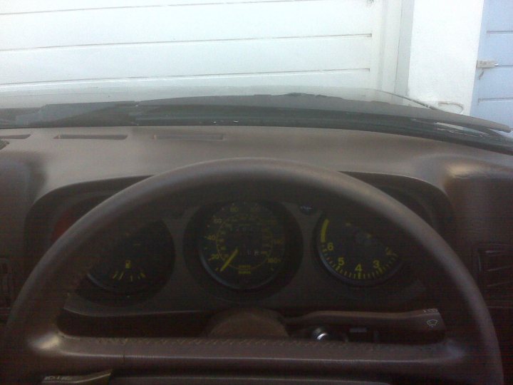 Love Pistonheads Hate - The image shows a close-up view of the portion of a car's steering wheel and dashboard where the speedometer and another smaller gauge are prominently visible. The gauges display yellow lines on a black background, suggesting an easy way of judging speed. Behind the gauges, there is a glimpse of the car's interior, and through the windshield, the exterior of a house with a white door and a light blue door can be seen, indicating the possibility of being near a residential area. The focus of the image is primarily on the gauges and the steering wheel, with the car's interior details slightly out of focus.