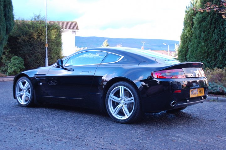 Pistonheads - The image shows the rear view of a black sports car in a driving position, parked on a tarmac driveway. The car's sleek design and polished finish reflect its sporty appeal. It features a spoiler on the back, flared wheel arches, and dual exhaust pipes. The setting suggests a residential area with greenery and a building visible, but the focus remains on the car itself.