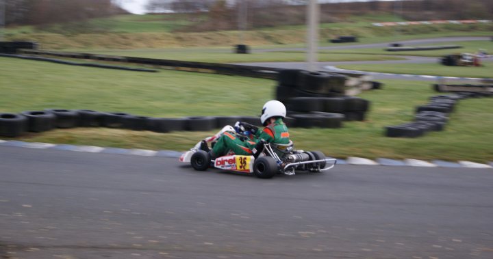 Trackday Pistonheads Action - The image showcases a thrilling scene of a man on a miniature race car tracked in black and white. The racer, donned in a green and white racing suit, is leaning to the left while navigating the track, which is lined with a mesh of black tires. The rear view mirrors of the race car suggest that the racer might be looking ahead, scanning the race course for potential opponents or dangers. The finish line of the track is visible at the background, adding to the excitement and anticipation of this competitive race.