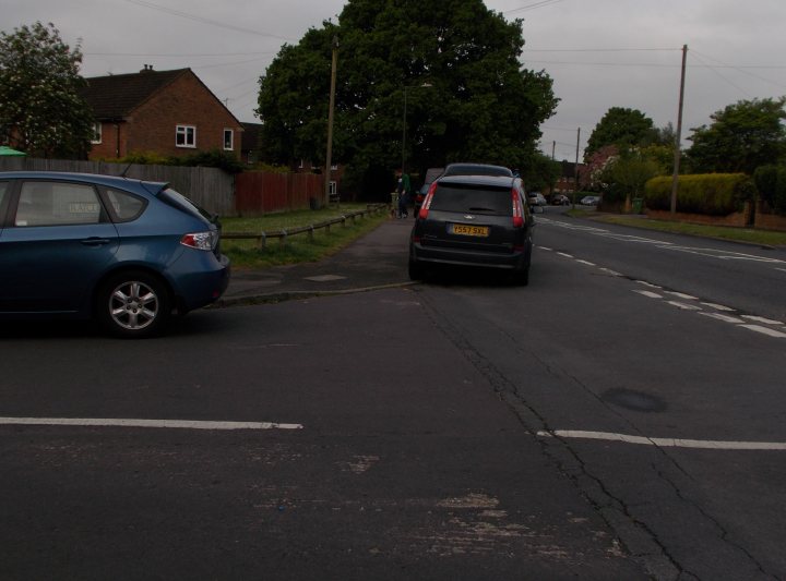 The BAD PARKING thread [vol3] - Page 324 - General Gassing - PistonHeads - The image is a photograph of a quiet street scene. In the foreground, a dark-colored car is parked to the right side of the image, and a blue car is parked directly opposite and is the vehicle closest to the viewer. The street appears to be in a residential area with some trees visible in the background. There are no people visible in the image. The sky is overcast, and the lighting suggests it could be early morning or late afternoon when the sun is low on the horizon. The image has a naturalistic style with colors that are typical of outdoor environments.