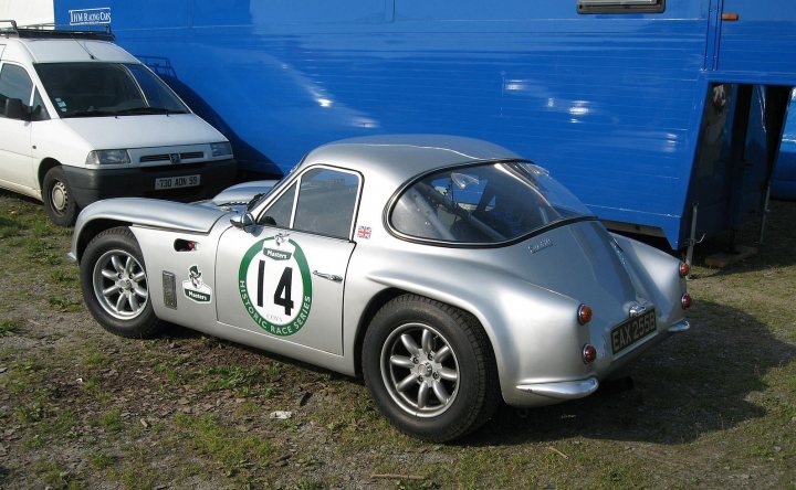 Early TVR Pictures - Page 159 - Classics - PistonHeads - The image features a shiny, silver sports car parked on a gravel surface. It is an old Model 14, as indicated by a logo and the number 14 visible on its side. The sports car is situated alongside a blue trailer, and a white van is visible in the background. The scene is outdoors and the ground appears to be uneven due to the presence of gravel. The overall atmosphere of the image suggests a casual parking setting, likely in a private or semi-private space.