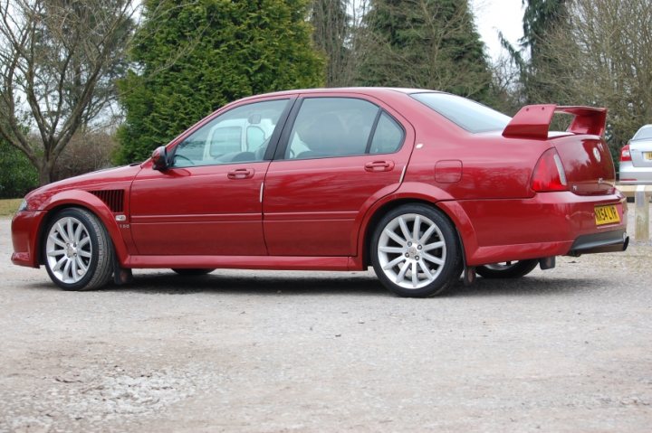 MG ZS 120 - Page 1 - Readers' Cars - PistonHeads - The image features a vibrant red sports car parked on a gravel surface. The car is adorned with a large rear spoiler, adding to its sporty appearance. The license plate of the car is clearly visible and reads "PH14 BK". The car's design and color make it the focal point of the image, while the trunk is open, hinting at recent or ongoing activity. The setting appears to be in a rural or semi-rural area, as indicated by the presence of trees and a tree stump in the background.