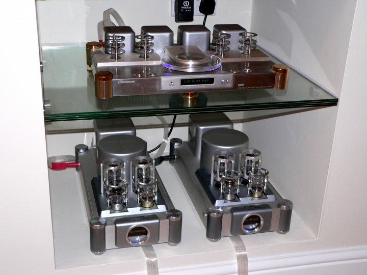 Set Pistonheads Hifi - The image shows a corner of a wall within which is mounted a glass shelf. On the shelf, there appears to be a piece of high end audio equipment, possibly a phono preamplifier or a similar device, characterized by multiple knobs and buttons. Beneath the shelf, also integrated into the wall, are two towering speaker amps or audio processors with a metallic finish. These display multiple control knobs and rows of smaller dials. The shelf and equipment seem to suggest a setup for playing vinyl records, given the appearance of a record player or phono cartridge connector on top of the shelf. The overall style of the equipment suggests a focus on quality and precision.