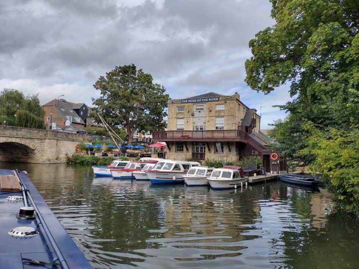 The canal / narrowboat thread. - Page 34 - Boats, Planes & Trains - PistonHeads UK - The image depicts a serene river scene. There are several boats docked along the riverbank, their vibrant colors adding life to the tranquil water. A small bridge arches over the river, providing a passage for pedestrians or vehicles. Buildings can be seen in the background, indicating that this peaceful scene is located within an urban area. The overall atmosphere of the image suggests a calm and leisurely setting, perhaps a popular spot for locals or tourists to enjoy a boat ride or simply take in the view.