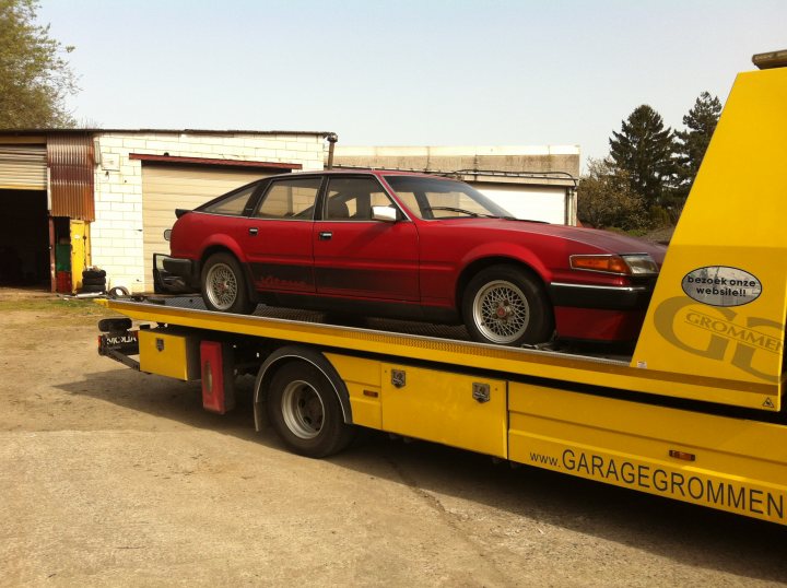 Pictures of my "NEW" SD1 Vitesse - Page 1 - Rover - PistonHeads - The image shows a vintage red car loaded onto a yellow tow truck. The car has a black interior, and it's positioned in the tow truck bed, ready to be transported. In the background, there is a white building with a wooden door and a large tree. In the foreground, there's some debris on the ground, but the focus remains on the car and the tow truck.