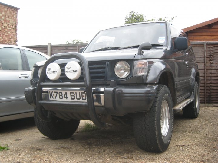 Offroader Cheap Pistonheads - The image shows a black SUV parked on a driveway. It has a distinctive bumper grille that is textured, with a 'Bud' logo on the right side. The vehicle is positioned in front of a wooden fence, and part of another vehicle is visible to the left. The sky is overcast, suggesting an overcast or gloomy day. The ground appears to be covered in a layer of dry leaves, indicating it might be autumn or the area is prone to falling leaves.