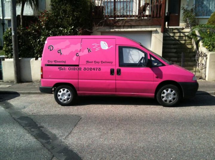 Goodness Pistonheads Sake - The image features a small pink van parked on a street. The van is vibrant and appears to be used for commercial purposes, as evidenced by the phone number displayed on the side doors. On the window of the van, there is a playful, hand-drawn depiction of a smirking face that seems out of place given its child-like appearance. The street on which the van is parked is residential and blends into the background of the image.