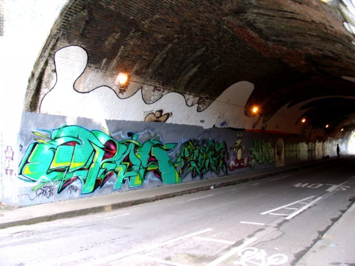 Pistonheads Tunnel Bristol - The image depicts a stark, urban landscape. A concrete tunnel, serving as an underpass for a road, is adorned with vibrant graffiti. The graffiti tag "DK" is prominently displayed, overlapping with markings in different colors like green and red. The lighting within the tunnel is dim, emphasizing the boundary between the cityscape and this artistic haven.