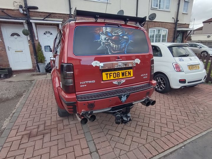 A car is parked next to a parking meter - The image shows a red SUV parked on the side of a brick road. The car is adorned with an array of accessories and modifications, including a roof rack, exhaust pipes, and a body kit that gives it a sporty appearance. There's also a large decal or sticker on the back window featuring an image of what appears to be a creature or monster. The vehicle has its rear door open, revealing a spare tire mounted under the hatchback. Behind the car is a house with a white door, and there are other cars parked in the background. A yellow license plate is visible on the car.