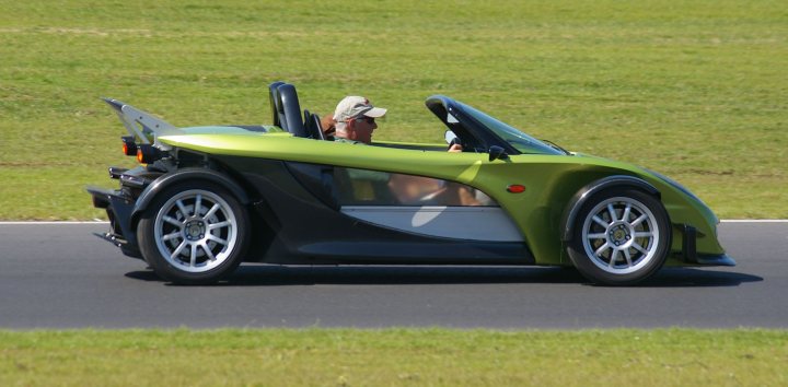 Snetterton Lotus Festival 25/26 June - Page 1 - General Lotus Stuff - PistonHeads - The image captures a dynamic scene on a green grass track. Dominating the center of the frame is a sleek, green three-wheeled car. The car appears to be in motion, with the front wheel turned slightly to the left. Two individuals are aboard the car, with the driver at the front and the passenger behind. Both appear to be donning helmets for safety, suggesting that they might be participating in a driving event on the track. The tire tread indicates the car's readiness for handling the grass track surface. The background reveals the open expanse of the grass track, stretching out to meet the horizon in the distance.