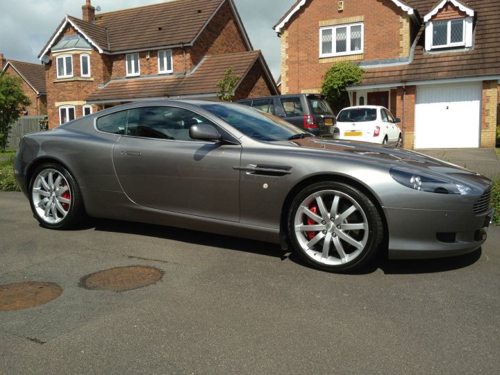 New "baby" arrived this weekend - Page 1 - Northamptonshire - PistonHeads - The image features a new gray Aston Martin car parked on the driveway of a two-story residential house. The house has a brick facade, with the main feature being a prominent bay window. The car is sleek with a shiny exterior reflecting its contemporary design. In the background, there are other cars parked, and the scene appears to be on a clear day with good lighting.