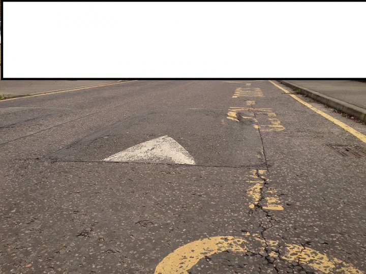 Gallardo's & road humps - Page 1 - Gallardo/Huracan - PistonHeads - The image depicts a quiet suburban street at an intersection with a triangular pothole. The street has a paved surface and the divisions between alternate sides of the road are marked by double yellow lines. There are no visible markings or traffic signs within the frame of the photo. The overall atmosphere is calm and unoccupied, indicative of stillness or a quiet moment outside of peak traffic hours.