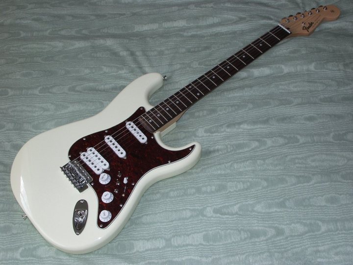 Pistonheads Luthiers - The image shows a white electric guitar with red trim on the neck. It appears to be a full-sized model, and the neck features a pickguard. The presence of pickups and the quality of the detail suggest it is a musical instrument. The guitar is lying on a fabric surface, which might be a cloth or a sheet. The focus is on the guitar, which is centrally placed in the frame, and there is no significant action or movement occurring, giving the image a static quality.