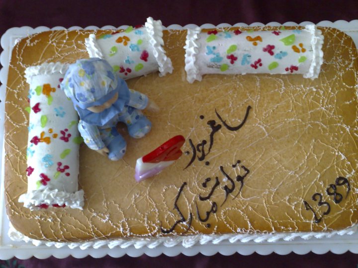 The image presents a colorful celebration featuring a square-shaped birthday cake. The cake has a foundation of brown wood-grain with white frosting trim, enhancing its festive look. Atop the cake, a blue toy robot stands next to a red toy train car, both adding an element of play to the scene. The cake is adorned with a simple greeting in Arabic, along with the number 4389, possibly indicating an important date or code. The cake is placed on a white plate with a subtle scalloped edge, placed on the right side of the image.