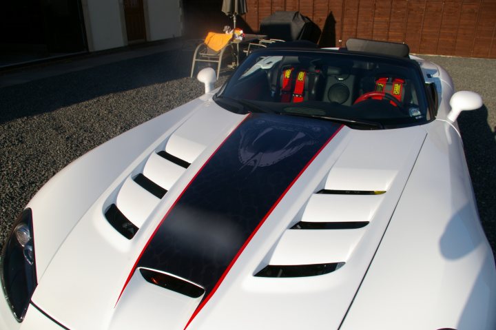 Blown But Not Blown - Page 1 - Vipers - PistonHeads - The image shows a white sports car parked on a surface that appears to be gravel or pebble. The vehicle has distinctive design features including a narrow front curve and a large spoiler with aerodynamic air ducts. There's a partial side view, revealing a helicopter logo adorning the car's side. The interior can be seen through the partially open top, showing red race seats. The background is an outdoor area with darker surfaces illuminated from the right side of the image, suggesting natural light or late afternoon sun.