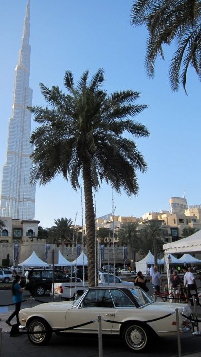 Anybody up for a trip to the emirates classic car festival  - Page 1 - Middle East - PistonHeads - The image captures a moment on a city street, where an eye-catching yellow car is parked. The car, which appears to be a convertible, is parked on the street, parallel to the sidewalk. The backdrop is quite impressive, featuring the Burj Khalifa, the tallest building in the world, towering over the cityscape. It's a beautiful day with clear skies, adding to the vibrancy of the scene. There are people walking around, contributing to the lively urban atmosphere.