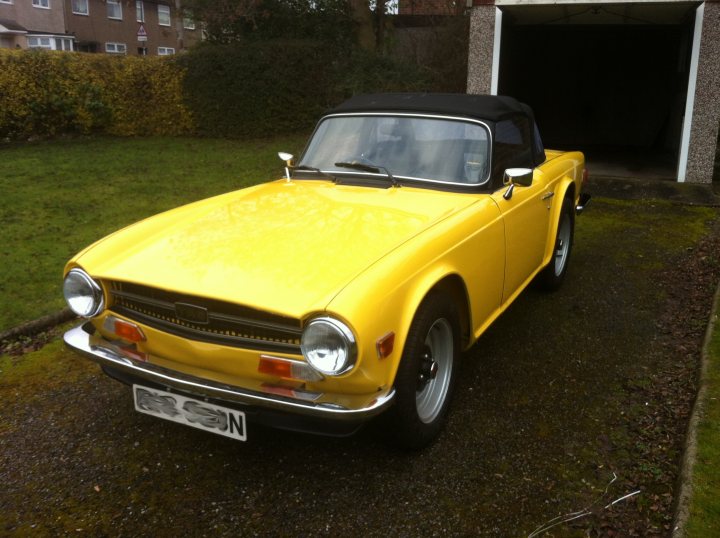 New Toy - TR6 - Page 1 - Triumph - PistonHeads - The image displays a yellow vintage sports car parked in a driveway. The car is compact and has a distinct design characteristic of vintage models. There's glass placed on the windshield, and a black fabric car cover is present. The background shows a residential area with a house and lush green grass. The setting appears to be under an overcast sky.