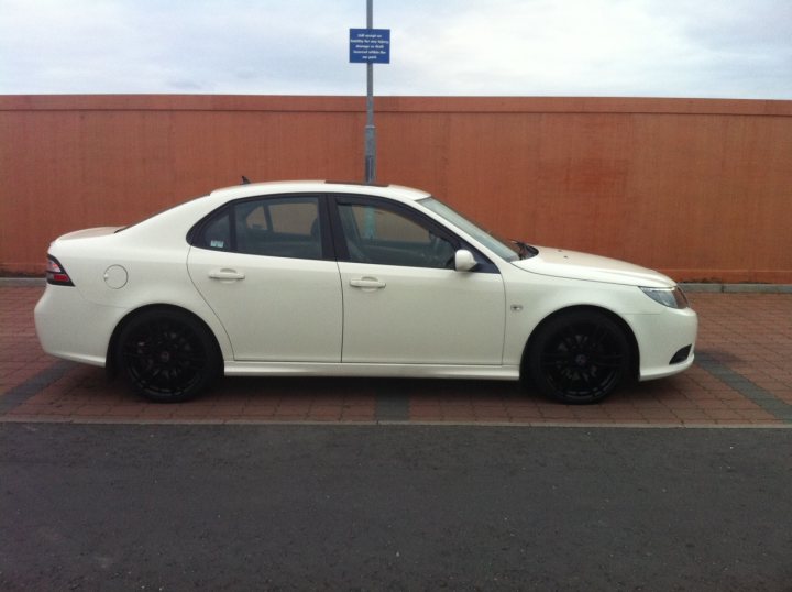 Show us your Saab!! - Page 14 - Saab - PistonHeads - The image showcases a white sedan parked on a brick surface against a partially visible orange wall. The car's black wheels contrast sharply with the vehicle's overall color scheme. Behind the car, there's a parking sign attached to a pole, hinting at the designated parking area. The sky appears overcast, suggesting cloudy weather conditions.