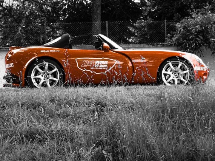 Show us ya cars... What you blasting around in 2015? - Page 7 - Yorkshire - PistonHeads - In the black and white photo, a vividly red Porsche 911 is the centerpiece, parked on a grassy area with weeds in the foreground. The car's sleek design and shiny details stand out against the contrasted background. The year model "24 Heures du Mans" and a racing event "Mille Miglia" are visible in the photo, suggesting that the car may have participated in these prestigious races. The car is facing away from the camera, providing a view of the black side-view mirrors and the textured hood. The day is overcast, casting even lighting over the scene.