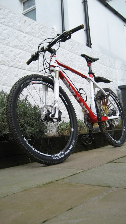 The "Show off your bike" thread! - Page 342 - Pedal Powered - PistonHeads - The image showcases a red and white mountain bike propped against a sturdy, white brick wall. The bike is equipped with a pair of semi-slick tires, suitable for both trail and road conditions. It features a suspension fork, enhancing its off-road capabilities, and a polycarbonate bottle carrier attached to the frame, providing a practical solution for carrying drinks on long rides. The bike is captured from a slightly elevated angle, giving a clear view of its design and features.