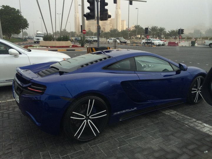 Middle East spotted thread - Page 63 - Middle East - PistonHeads - The image displays a vibrant blue sports car parked on a city street. The car is positioned at a traffic light on the right side of the street, which is painted in the typical red color. Various buildings can be seen in the background, indicating an urban environment. The traffic light features multiple lanes of traffic and a few other cars are visible in the distance. The car is at rest, with no immediate traffic visible.