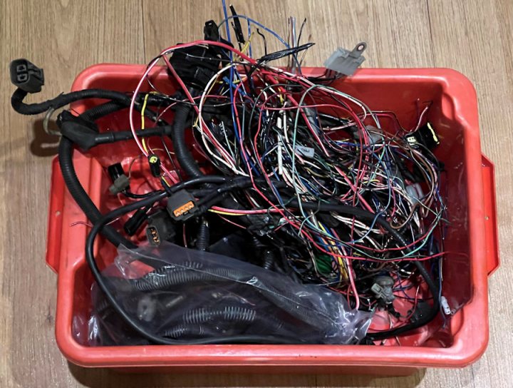 Pistonheads - The image shows an open red plastic container filled with a variety of colorful wires and connectors. These parts appear to be from electronic equipment, possibly circuit boards or harnesses. There are different types of plugs, coils, and insulated wires in various colors. A white tag is attached to the side of the container, although the text on it is not visible. The background suggests an indoor setting with a light-colored floor, but no distinctive features can be identified from this angle.