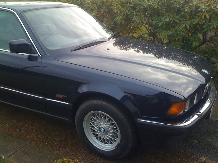 Shed Week Pistonheads - The image showcases a classic BMW car, parked on the street. The car is predominantly dark blue, with some light blue details adding to its vintage aesthetic. Its black top makes it look even more elegant. The car is positioned in front of a bush, which adds a touch of green to the scene. Despite the car looking polished, there is a small dent on the hood, which reveals beneath a yellow interior.