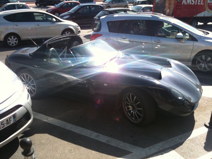 Pistonheads - The image shows a parking lot with several cars. The main focus is a convertible car with its top down, allowing sunlight to refract through the windshield onto the hood, creating a visually striking effect. The convertible car appears to be a two-seater, and it's parked next to a grey vehicle and in front of a truck. The photograph captures the vibrant interplay between light and reflection, emphasizing the design features of the convertible car. There are no people visible in the image. The style of the image is a straightforward, everyday photograph without any visible artistic manipulation.