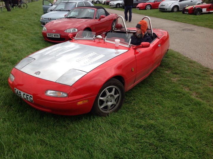 Ever seen a three wheeler MX5? - Page 1 - Mazda MX5/Eunos/Miata - PistonHeads - The image captures a vintage scene of a red convertible sports car parked in a lush, grassy field. The car, which is the main subject, has a convertible top down, revealing the interior. Parked next to it is another car, this one a white convertible. Both cars are parked defensively, ready to leave at a moment's notice, with the wheels turned towards the camera. A solitary figure, dressed in warm clothing, is standing next to the red car, possibly the owner or a passerby. In the backdrop, a row of other cars can be seen, their presence adding depth to the scene.