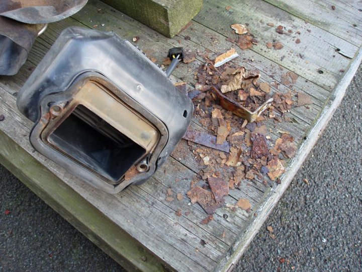 Dead Fan Heater Pistonheads - The image shows a close-up view of debris surrounding a platform. The debris is a mix of broken concrete, rusted metallic parts, and small stones. An unmistakable object that appears to be a cup or mug is forged on the concrete, with its brownish tint contrasting against the grey of the concrete and metal pieces. The platform itself is made of wooden planks. The ground beneath the platform is underlaid with dark-colored asphalt. The overall impression is that of a neglected or discarded area, with a somewhat melancholic or forgotten aesthetic.