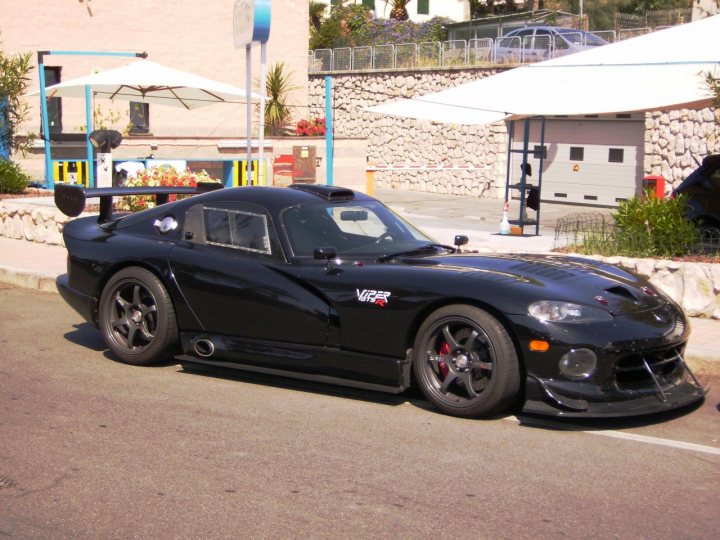 Supercars spotted, some rarities (Vol 4) - Page 23 - General Gassing - PistonHeads - The image shows a black coupe with a large rear wing parked near a storefront. The car is positioned on a street next to a sidewalk. In the background, there are various objects including plants, flower pots, and part of a building with a metal shutter covering a window. The ground is not fully visible, but it appears to be an asphalt surface. Given the context and items in the background, it's likely a commercial or residential area.