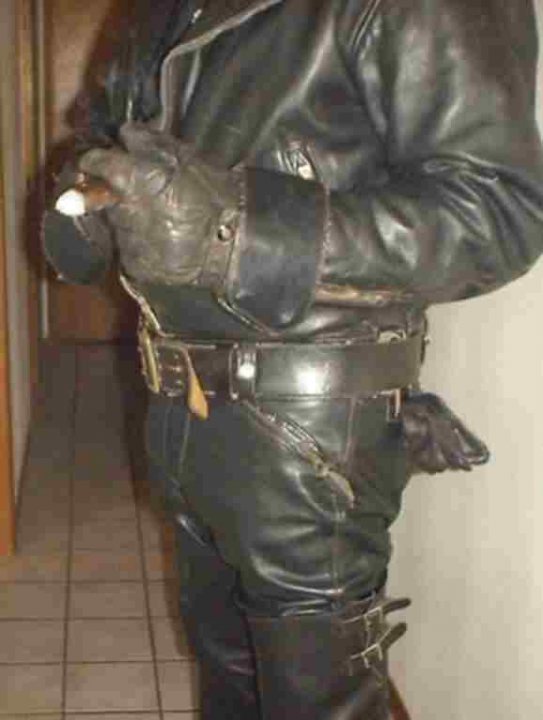 Jiggyziggy2 - The image shows the back of a person dressed in a leather outfit, which includes a jacket and what appears to be a girdle or a tool belt. The person has one hand on their hip and is holding an object in their other hand, although the object is not clearly visible. The individual is standing in an indoor setting, evident by the tiles on the floor. The leather attire suggests a texture contrast to the smooth tiles.