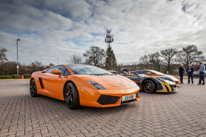 Supercar meet at Flying Horse PH clophill 22/3/15 - Page 9 - Herts, Beds, Bucks & Cambs - PistonHeads - The image captures an outdoor scene featuring a group of people and two sports cars. In the foreground, there's an orange sports car parked on a cobblestone surface, taking up a sizable portion of the image. Behind this orange car, there's another car positioned further back, appearing to be a green and black race car, which is smaller in comparison. 

Several people are standing around the area, some close to the cars and others slightly distanced. The vehicles seem to be the main focus of their attention. 

The background reveals a line of trees, adding a natural element to the scene. In the far distance, there is a utility tower, providing an urban touch to the image. 

The cobblestone surface on which the orange car is parked has a textured appearance, providing an interesting contrast to the smooth surfaces of the sports cars.