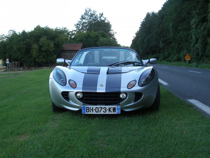 Going topless - Page 1 - Elise/Exige/Europa/340R - PistonHeads - The image shows a silver sports car parked on the side of the road next to a grassy area. The car has a prominent black and blue stripe along its body, suggesting it is a racing model or a special edition car. It is a bright and somewhat sunny day, and the car's reflective surface indicates it is likely in midday or early afternoon light. In the background, there are trees indicating a rural or suburban setting.