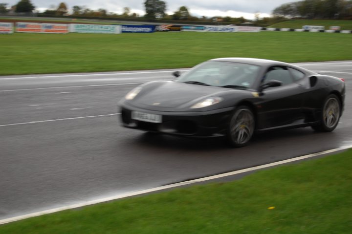 Stroke Association Day Supercar Pistonheads Castle Combe - The image portrays a sleek, black sports car speeding around a curve on a race track. The car is captured in a dynamic pose, leaning into the turn, emphasizing its agility and performance. The background is filled with race track infrastructure, including various advertising banners alongside the track. The sky above is cloudy, suggesting an overcast day, which might indicate cooler weather and better driving conditions.