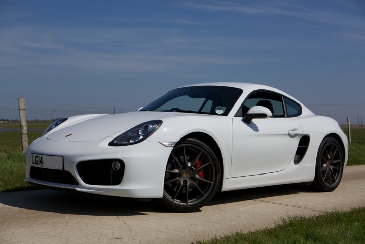 Boxster & Cayman Picture Thread - Page 12 - Boxster/Cayman - PistonHeads - The image presents a white Porsche sports car positioned on a grassy area adjacent to a concrete path. The car is parked, facing towards the viewer. It's a clear day with a blue sky, and in the background, there are wooden fences. The car's design includes aerodynamic curves and features sports elements such as a large spoiler and black wheels with red strips. The license plate is visible.