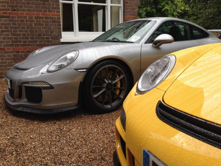 A red car is parked on the side of the road - Pistonheads - The image showcases two cars parked side-by-side on a driveway. The car on the left is a silver Porsche with a sleek design, while the car on the right appears to be a yellow vehicle with a distinctive front grille. Both cars have black wheels, but the Porsche has a smaller grille and silver wheels. The driveway has pebbles, giving a texture to the ground. There is a brick building in the background, suggesting a residential area.