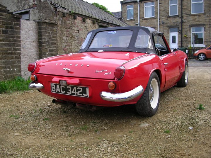 Driver Pistonheads Tallest - The image features an iconic red Volkswagen Beetle car parked in a gravel driveway. The car is positioned facing the right side of the frame, giving a view of its front, where the license plate reads "BAC 342 J." The Beetle has a sloped windshield and the distinguishing straight, chrome-covered fender. The driveway extends into the background, leads to a house, and is flanked by lush greenery. Plants are growing in the cracks of the driveway, adding a touch of nature to the scene.