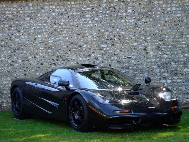 Moving Motor Show Pics  2011 - Page 1 - Goodwood Events - PistonHeads - The image depicts a black sports car parked on a grassy area. The car features smooth, aerodynamic curves that are characteristic of high-performance vehicles. It has a clear, glossy finish that reflects the sunlight, which is on the right side of the photo, creating beams of light around the car. The background shows a wall or fence, which is partially obscured by the vehicle, giving the impression that the car is parked relatively close to it. The setting appears to be outdoors during the day.