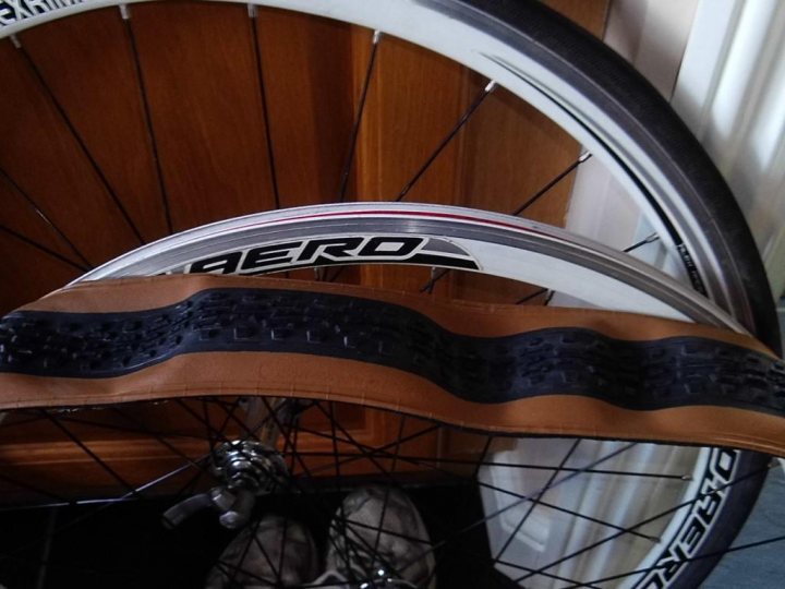 My new tyres are too tight for the rims!!! - Page 3 - Pedal Powered - PistonHeads - The image captures a scene from a garage or workshop with a focus on two bicycle wheels resting against a wooden door. The wheel in the foreground is a white-walled aero model with a distinctive color scheme of black, red, and white. The other wheel is a large Black RockShox model with its wheel guard removed to expose the spokes. The overall setting suggests a space where cycling equipment is stored or repaired.