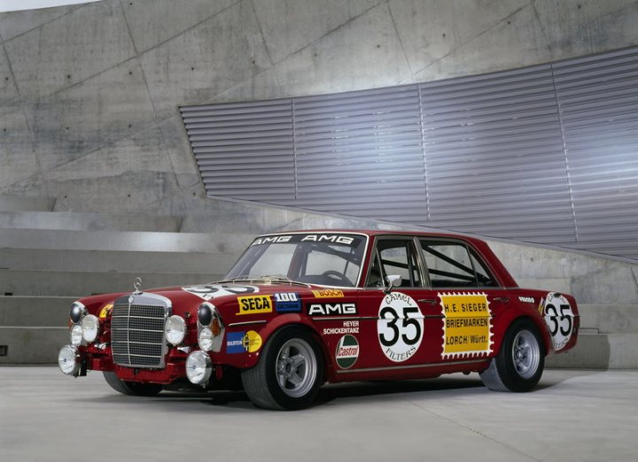 Pistonheads Masculine - The image presents an event where a vintage red car, possibly designed for racing, is prominently displayed. The car is parked in front of a modern assembly plant, suggesting a focal point on automotive heritage merging with contemporary technology. A prominent feature on the car is the "AMG" logo, indicating a high-performance brand, and there are other sponsor and performance-related brands and logos visible on the car's side. The environment is industrial, with concrete and metal structures providing a stark contrast to the car, highlighting the vehicle as the center of interest in this image.