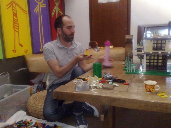 This image captures a playful moment in a room that seems to be a mix of a workshop and a living space. A bearded man is sitting on a chair, engrossed in the assembly of a Lego structure on the dining table. The table is filled with various Lego pieces and other toys, suggesting a child-friendly environment. On the table, there are pieces of stone jenga, a clue to a possible diverse collection of toys in this space. Behind the man, two large paintings adorn the wall, adding an artistic touch to the room. There's also a television in the background, indicating the room could be used for relaxation or entertainment too. The atmosphere seems creative and fun, with the man's focus on his Lego build.