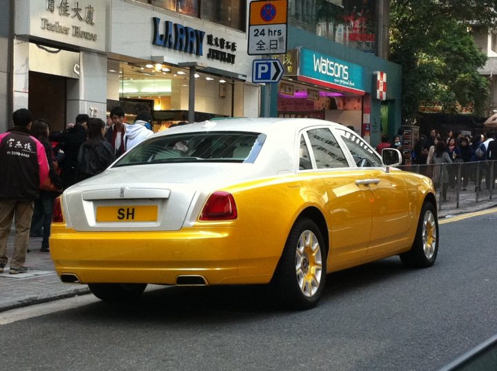 Pistonheads - The image shows a scene from a city, with a bright yellow car as the focal point. The car is stopped at a traffic light, which is currently red, indicating a halt in motion. The car is adorned with a license plate, reading "SH" and has the word "Larry" written on the side, hinting at personalization. The car is parked on the street, adjacent to a store with a sign that reads "Larry shopping mall" and a parking area with small potted plants overlooking the space. There are several people visible in the background, going about their day, adding a lively atmosphere to the urban setting.