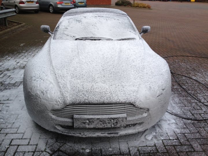 Back in the fold and its feeling good!!!! - Page 1 - Aston Martin - PistonHeads - This image shows a white car partially covered in snow, likely just parked, as evidenced by its shiny surface and visible glass. It appears to be in a parking lot with other vehicles and some green plants in the background. The bricks of the parking area have a frost or ice buildup, giving further indication of cold weather. The setting suggests a wintertime day in an urban environment.